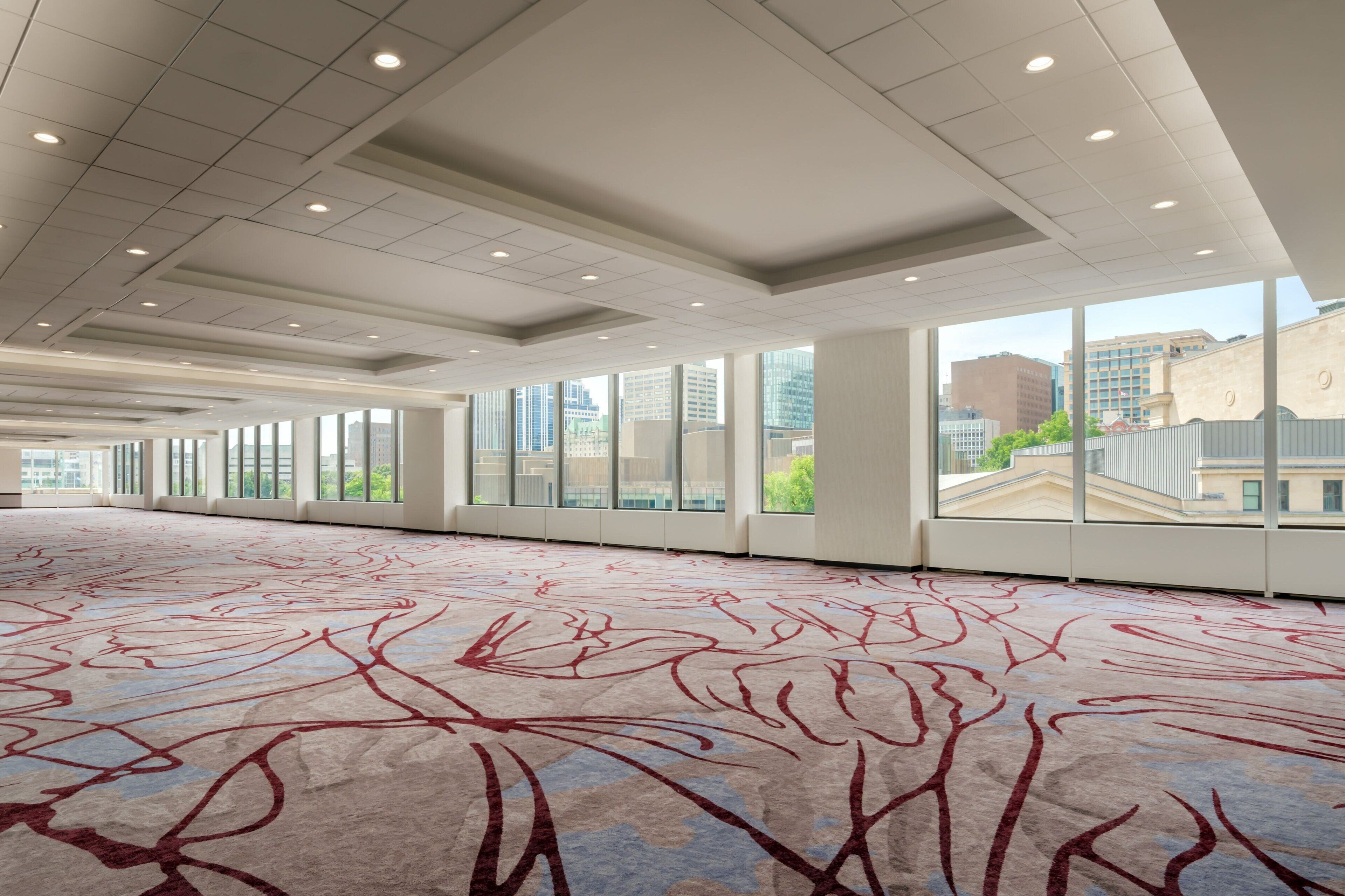 A spacious room with a large carpet featuring abstract red and blue patterns, large windows offering city views, and a modern ceiling with recessed lighting.