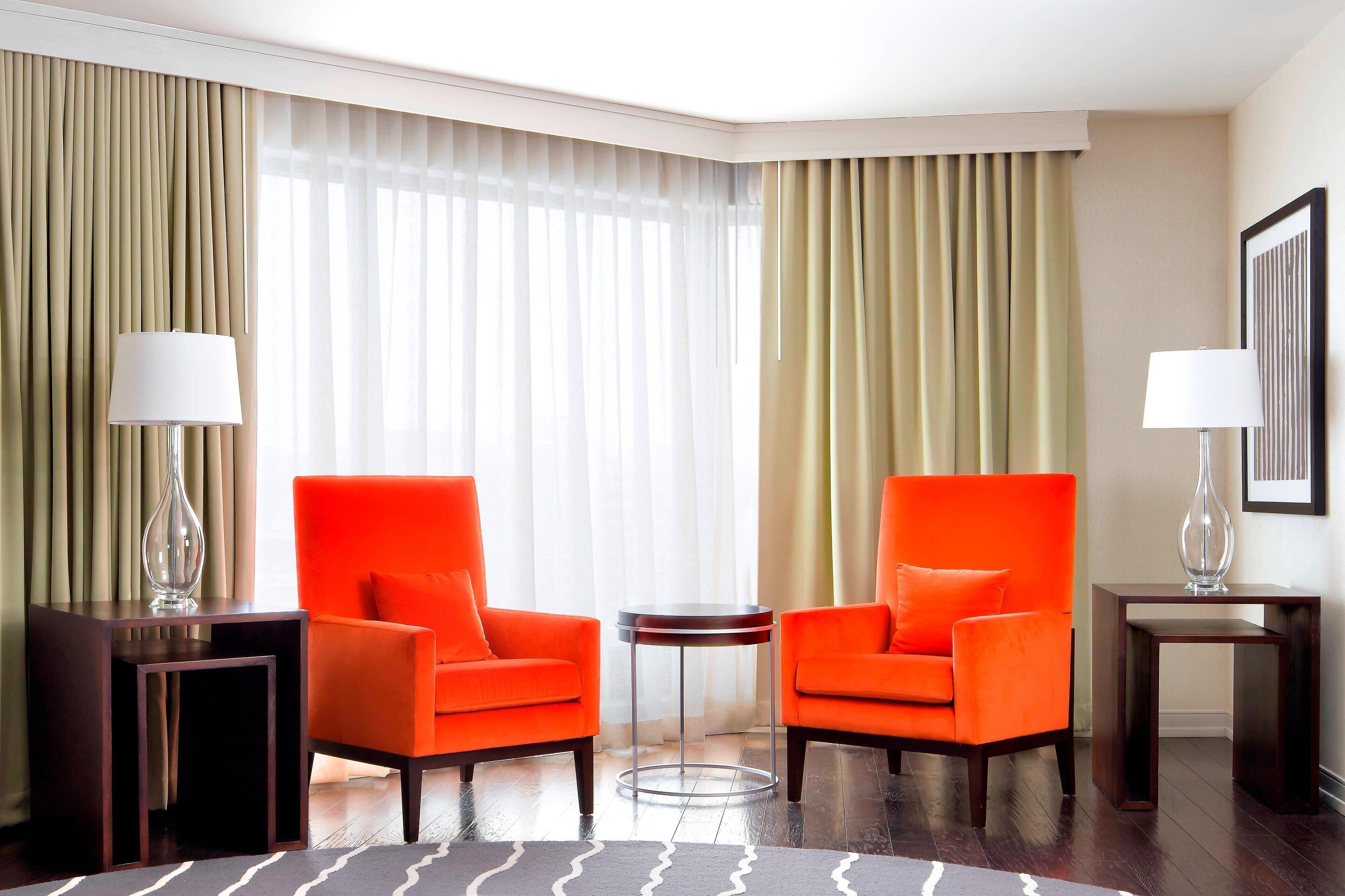 A hotel room with two orange armchairs, a small round table, and two lamps.
