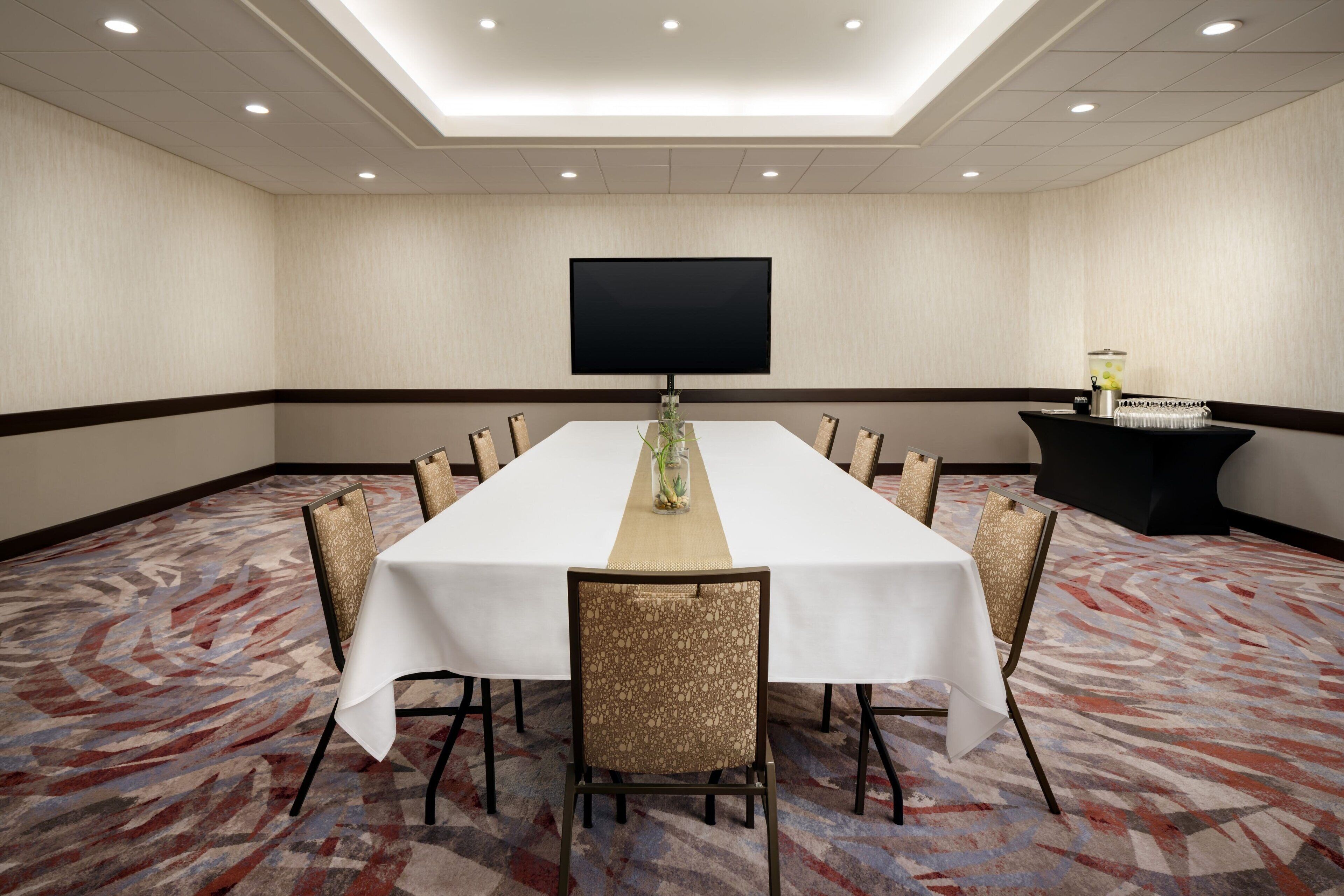 A conference room with a long table, chairs, a flat-screen TV, and a beverage station.