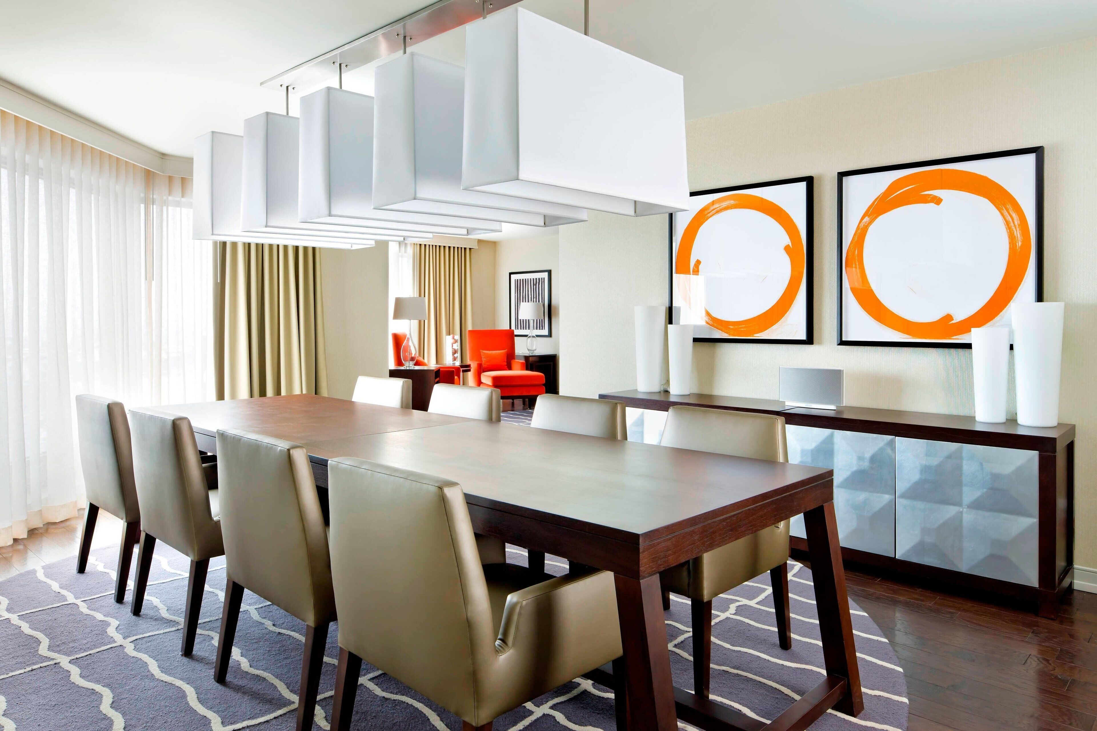 A hotel dining area with a long wooden table, beige chairs, a modern chandelier, and abstract artwork on the wall.