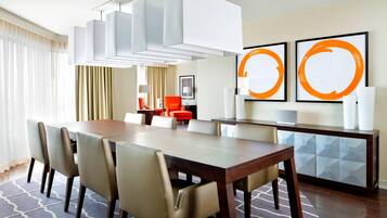 A hotel dining area with a long wooden table, beige chairs, a modern chandelier, and abstract artwork on the wall.