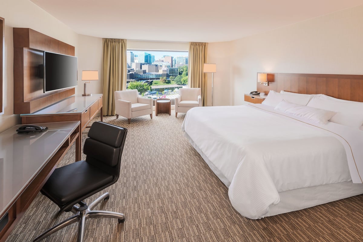 A hotel room with a large bed, a desk with a TV, two chairs, a small table, and a view of the cityscape.