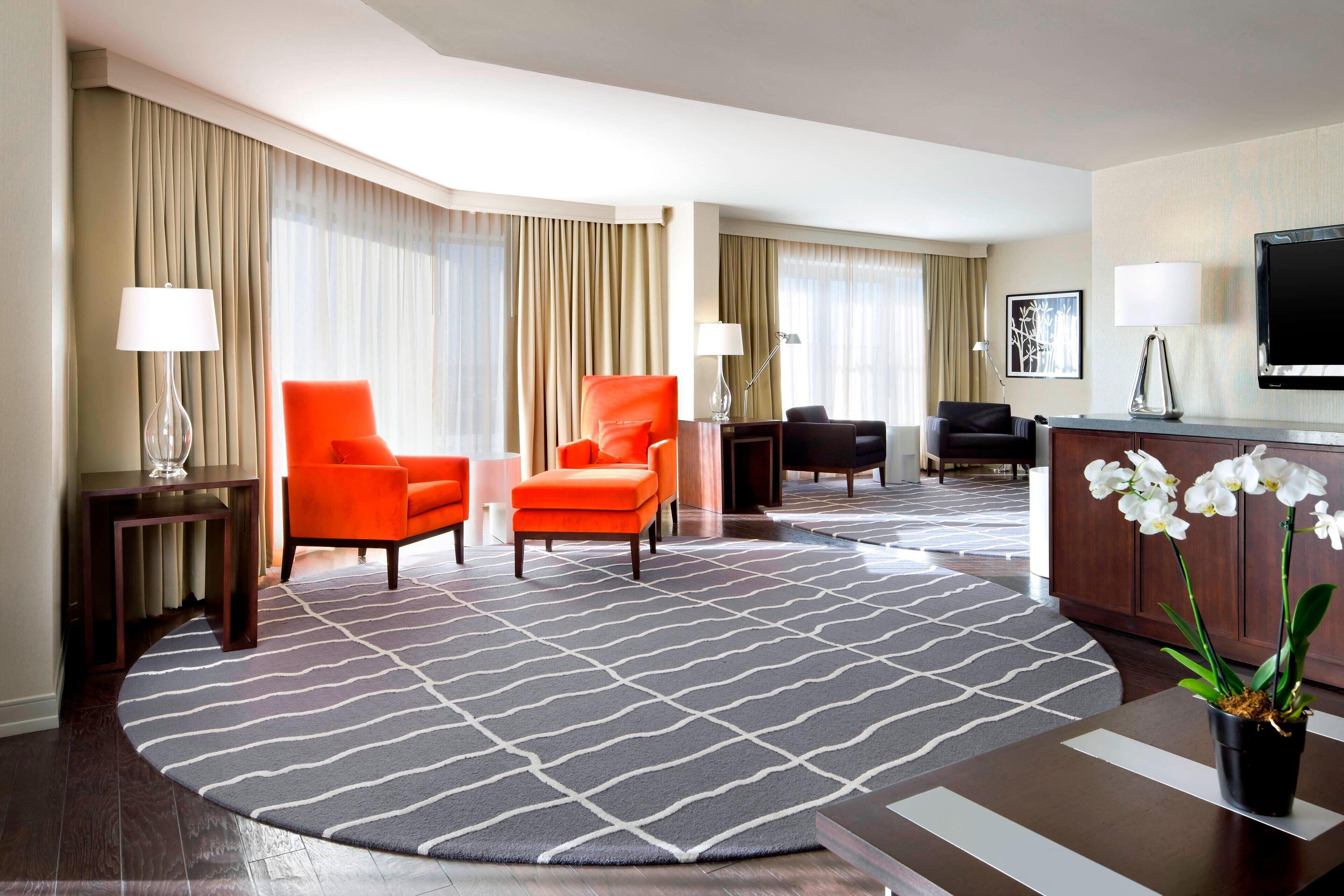 Habitación de hotel con una alfombra grande de estampado llamativo, dos sillones naranjas, un sofá negro, una mesa de centro de madera y un televisor montado en la pared.