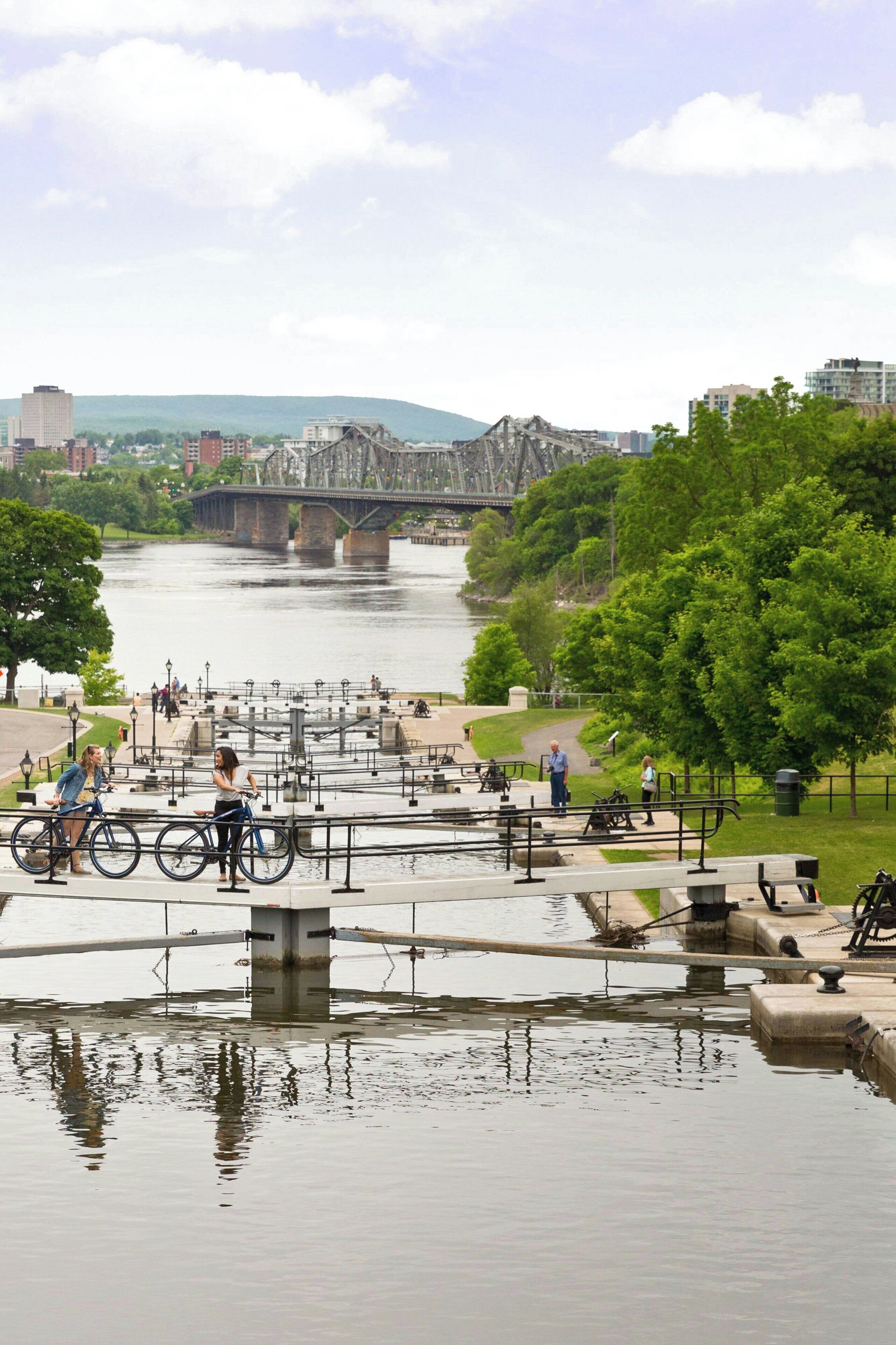 A riverside park with a bridge in the background, bike racks, and people walking.