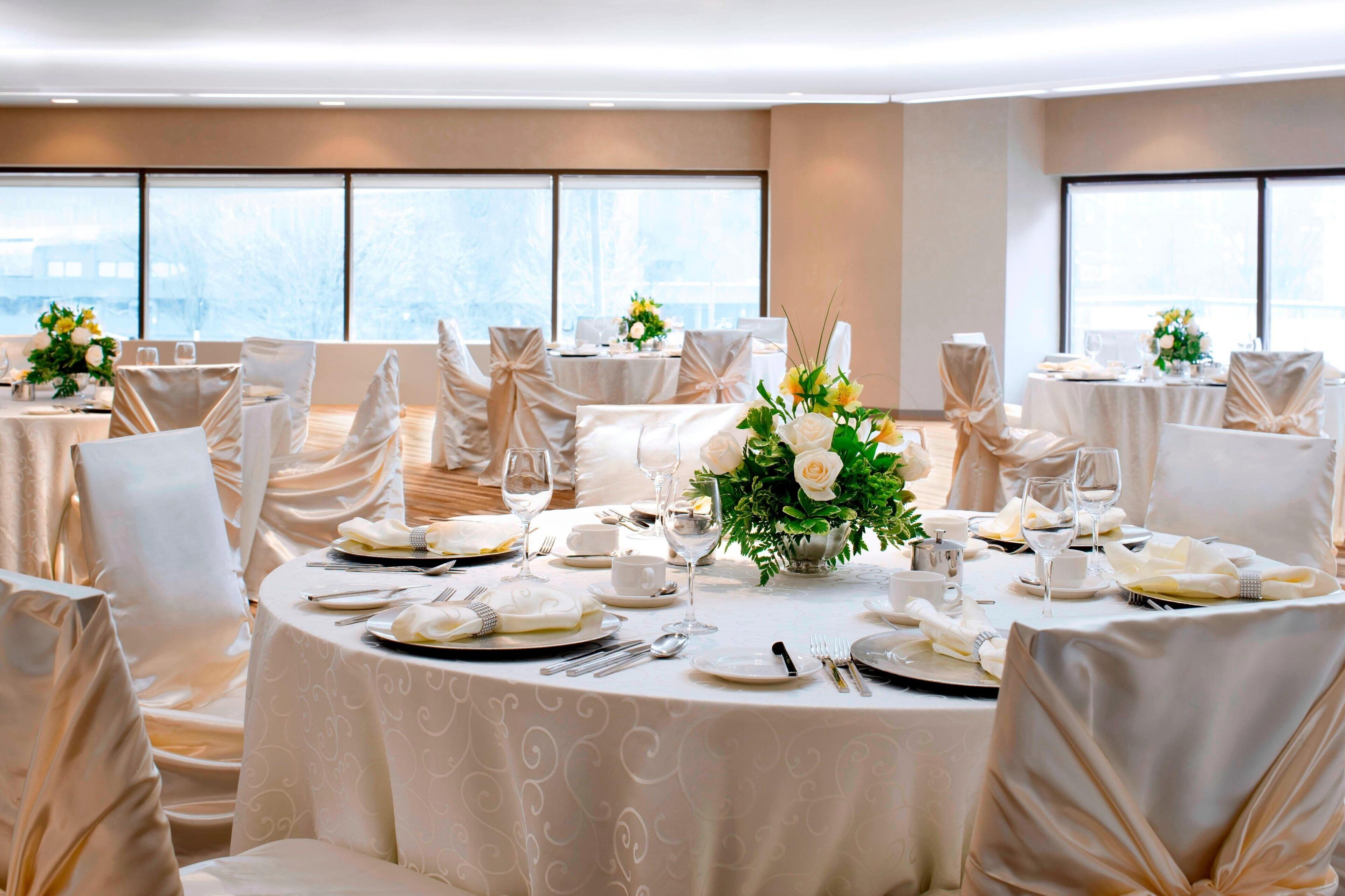 A banquet hall with round tables set for a formal event, featuring white tablecloths, chairs with white covers, and floral centerpieces.