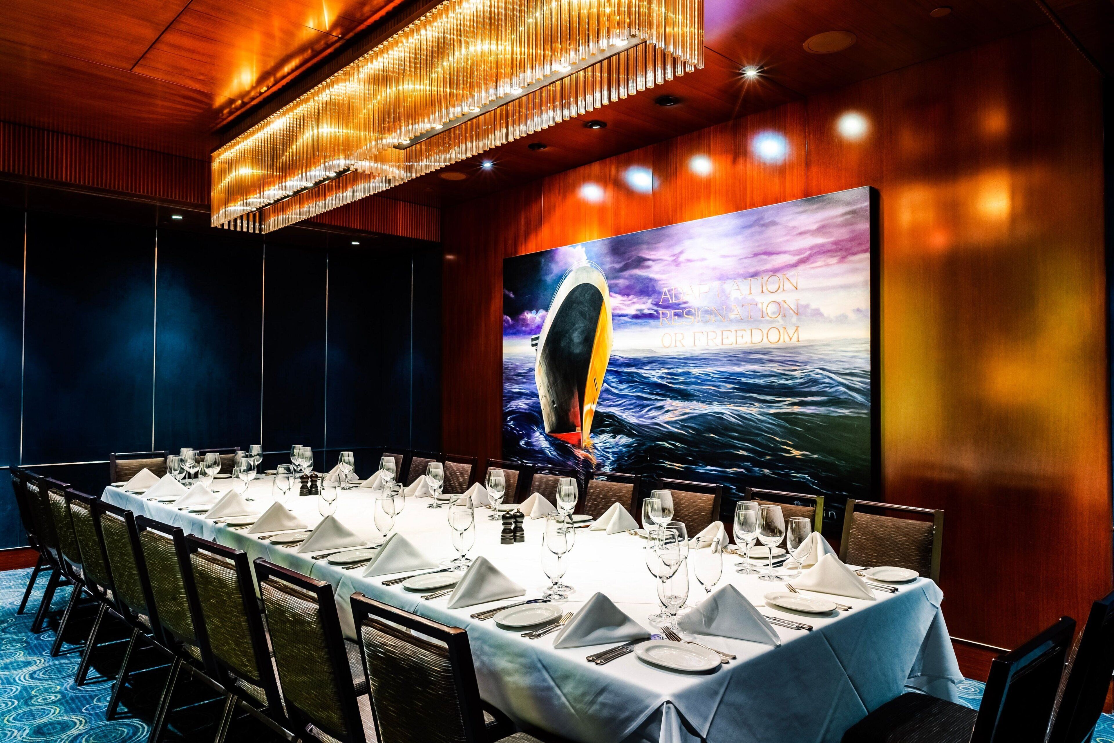 Une salle de conférence avec une longue table préparée pour une réunion, un grand écran diffusant une vue sur la mer, et un lustre au plafond.