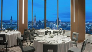 Un comedor con mesas redondas dispuestas para seis personas, amplias ventanas que ofrecen una vista de la ciudad al atardecer.