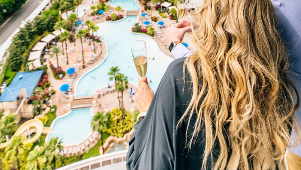 A man holding a glass of champagne overlooking a resort with a pool, lounge chairs, and a large building in the background. - Hyatt Regency Orlando (Orlando)