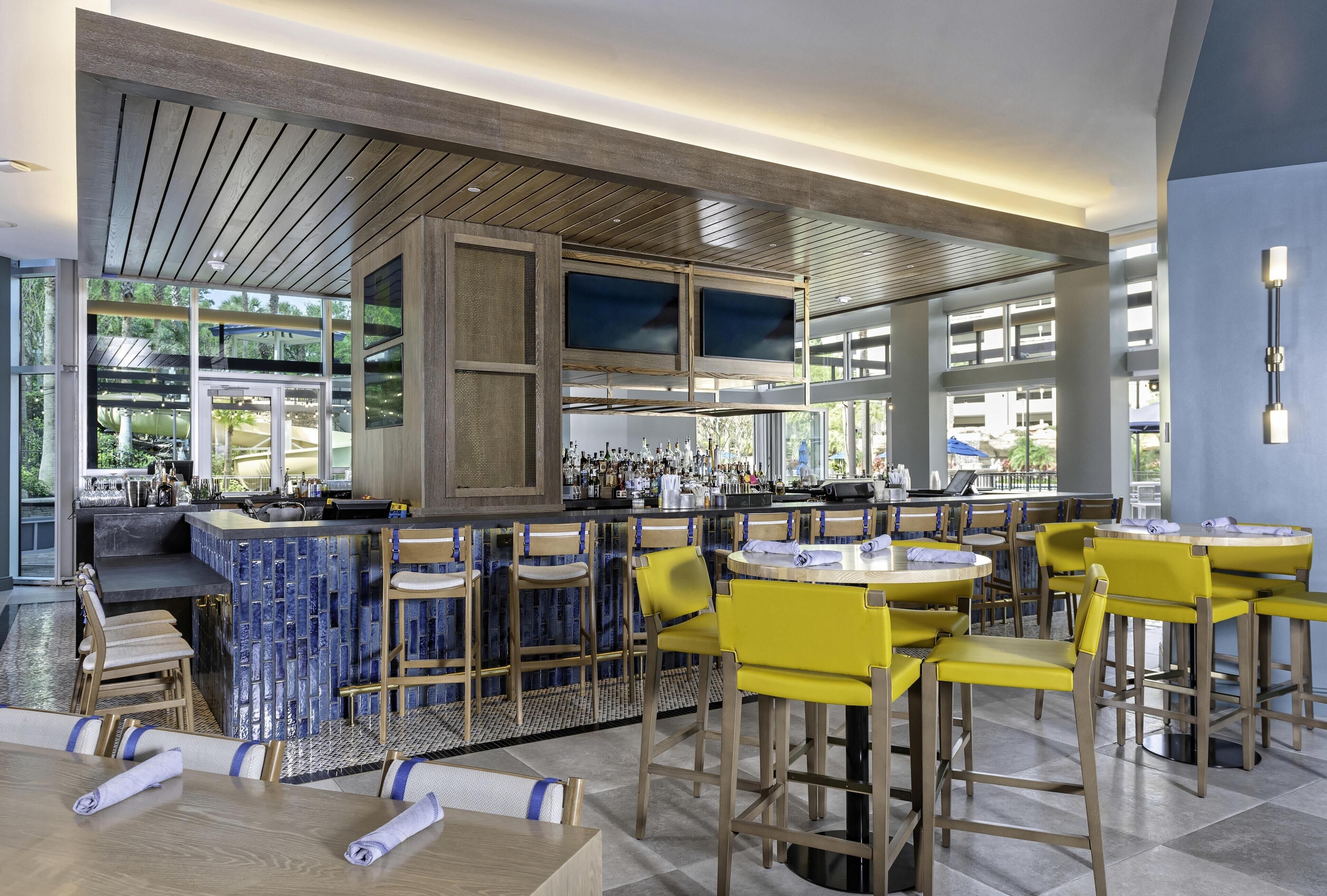 Un área de bar moderna con mostrador, taburetes y una zona de estar con mesas redondas y sillas.