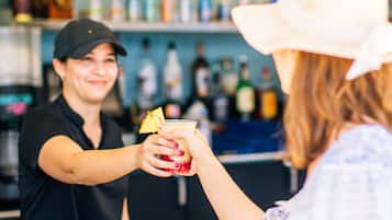 Eine Person hält ein Getränk mit Ananas-Garnitur an einer Theke, auf der ein Schild mit dem Schriftzug 'Welcome to Coconut's' steht.