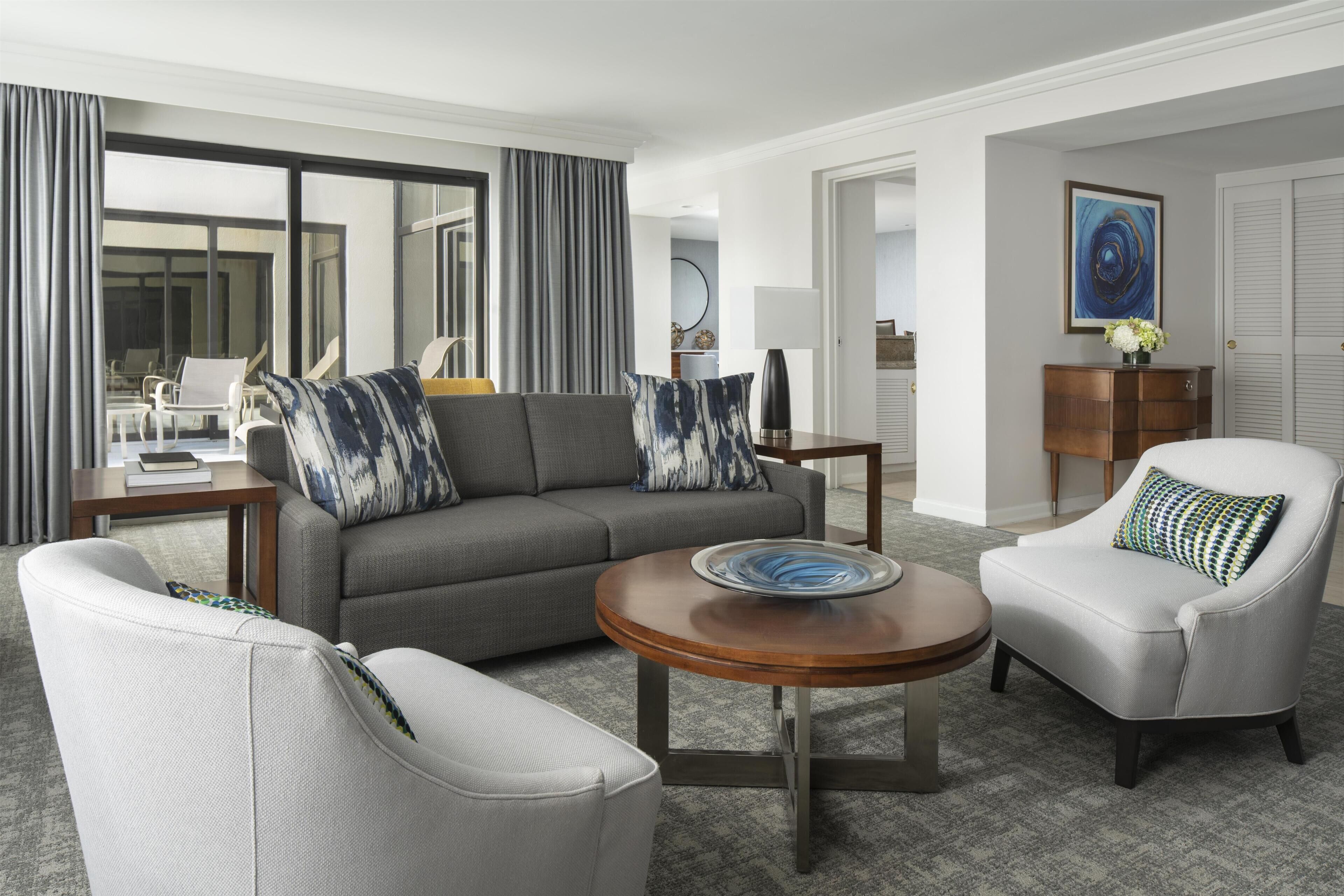 A modern hotel room with a gray sofa, white armchairs, a wooden coffee table, and a view of the outside.