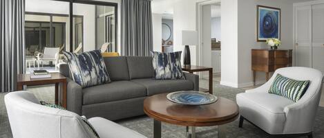 A modern hotel room with a gray sofa, white armchairs, a wooden coffee table, and a view of the outside.
