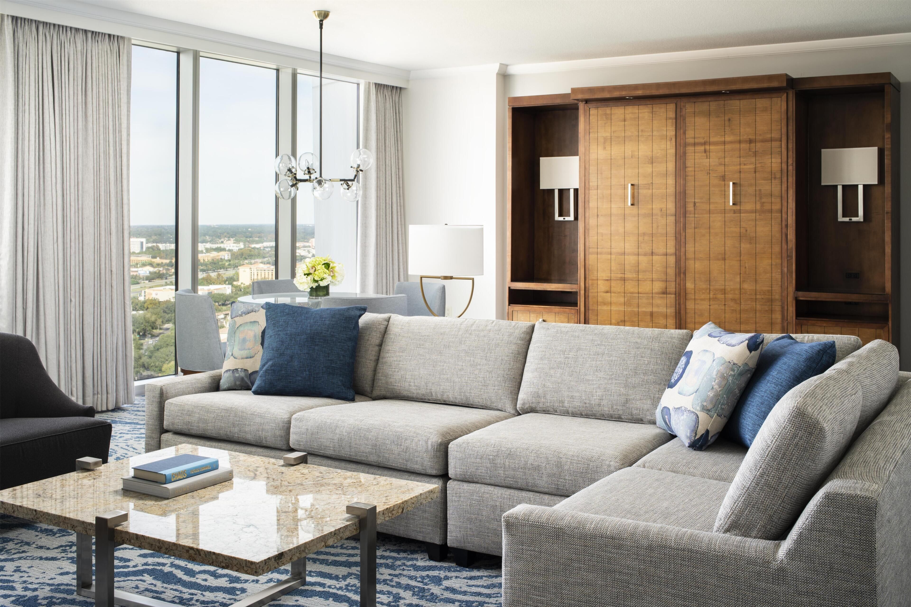 A modern hotel room with a sectional sofa, a coffee table, a large window with a city view, a wardrobe, and a chandelier.