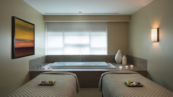 A room with a large bathtub, two beds, a painting, and a wall-mounted light.