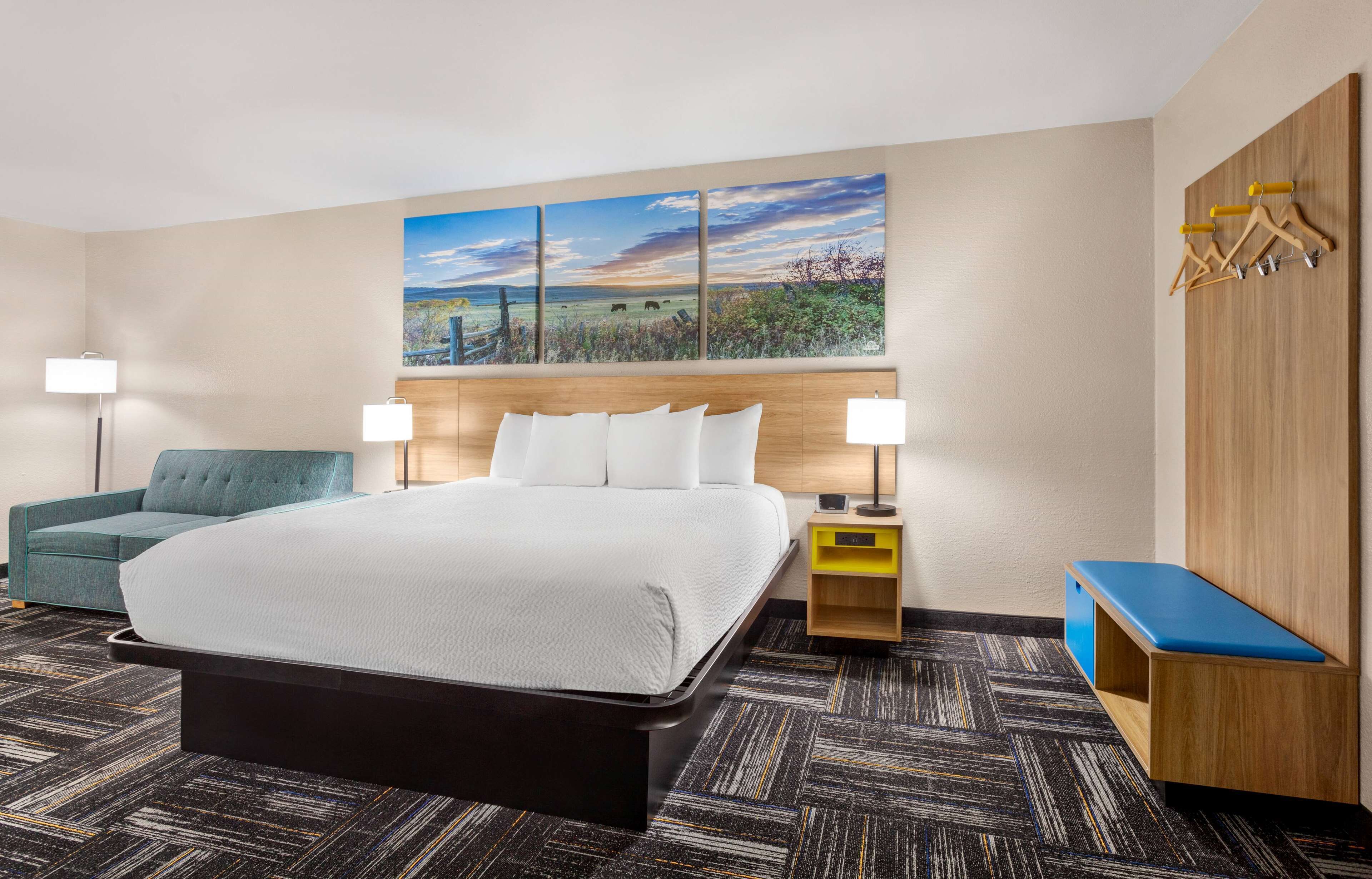 Quarto de hotel com uma cama grande, duas luminárias de cabeceira, um sofá azul, uma cabeceira de madeira, um guarda-roupa de madeira e um quadro com uma paisagem.