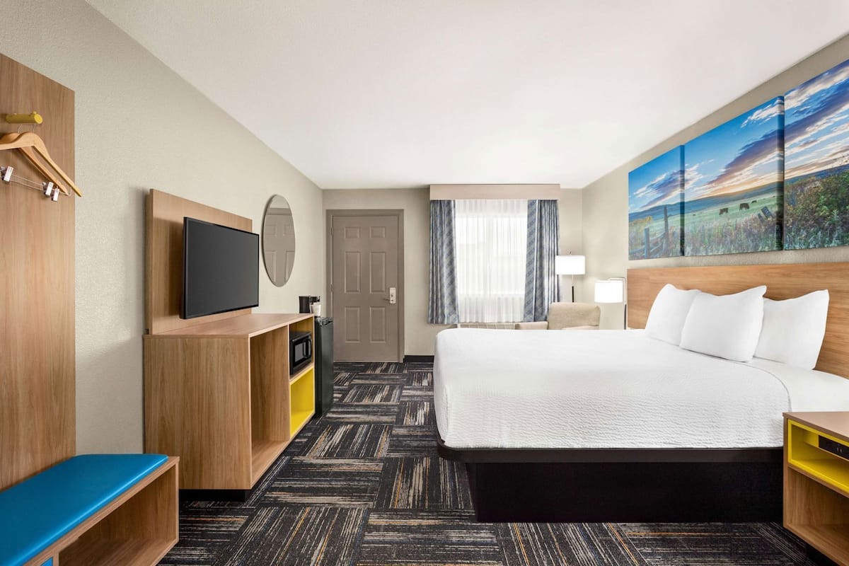 A hotel room with a bed, a TV, a desk, a mirror, a door, and a painting on the wall.