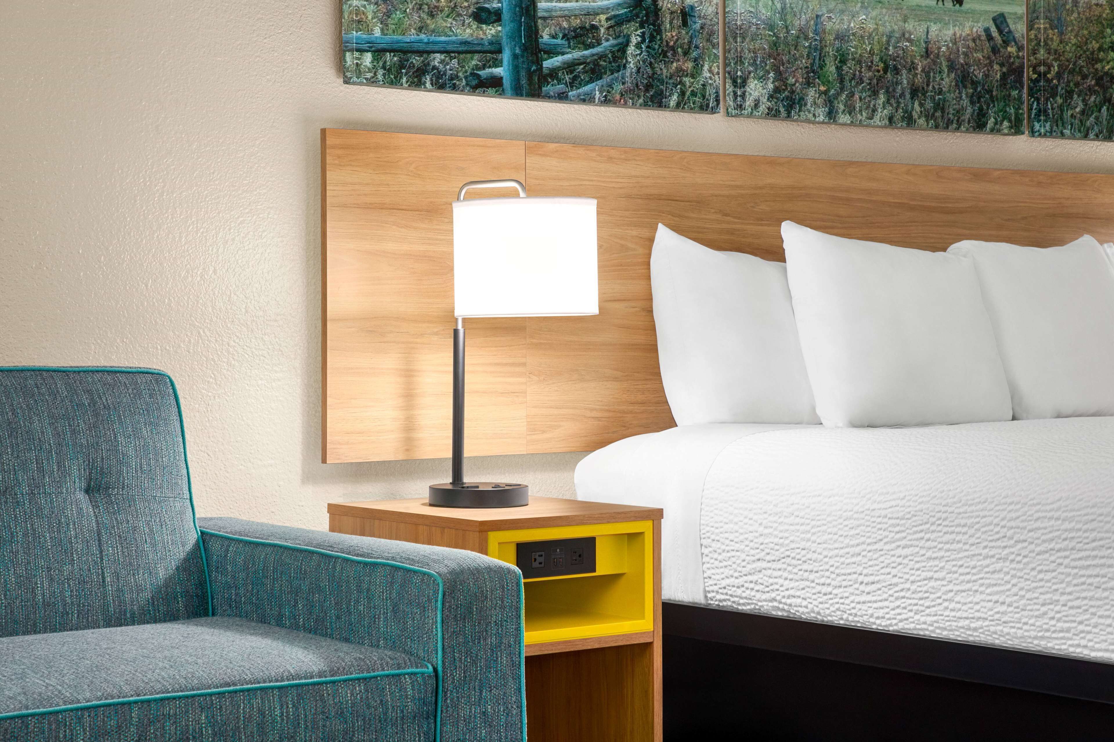 Habitación de hotel con cama, mesita de noche con lámpara, una silla y un cajón de almacenamiento amarillo.