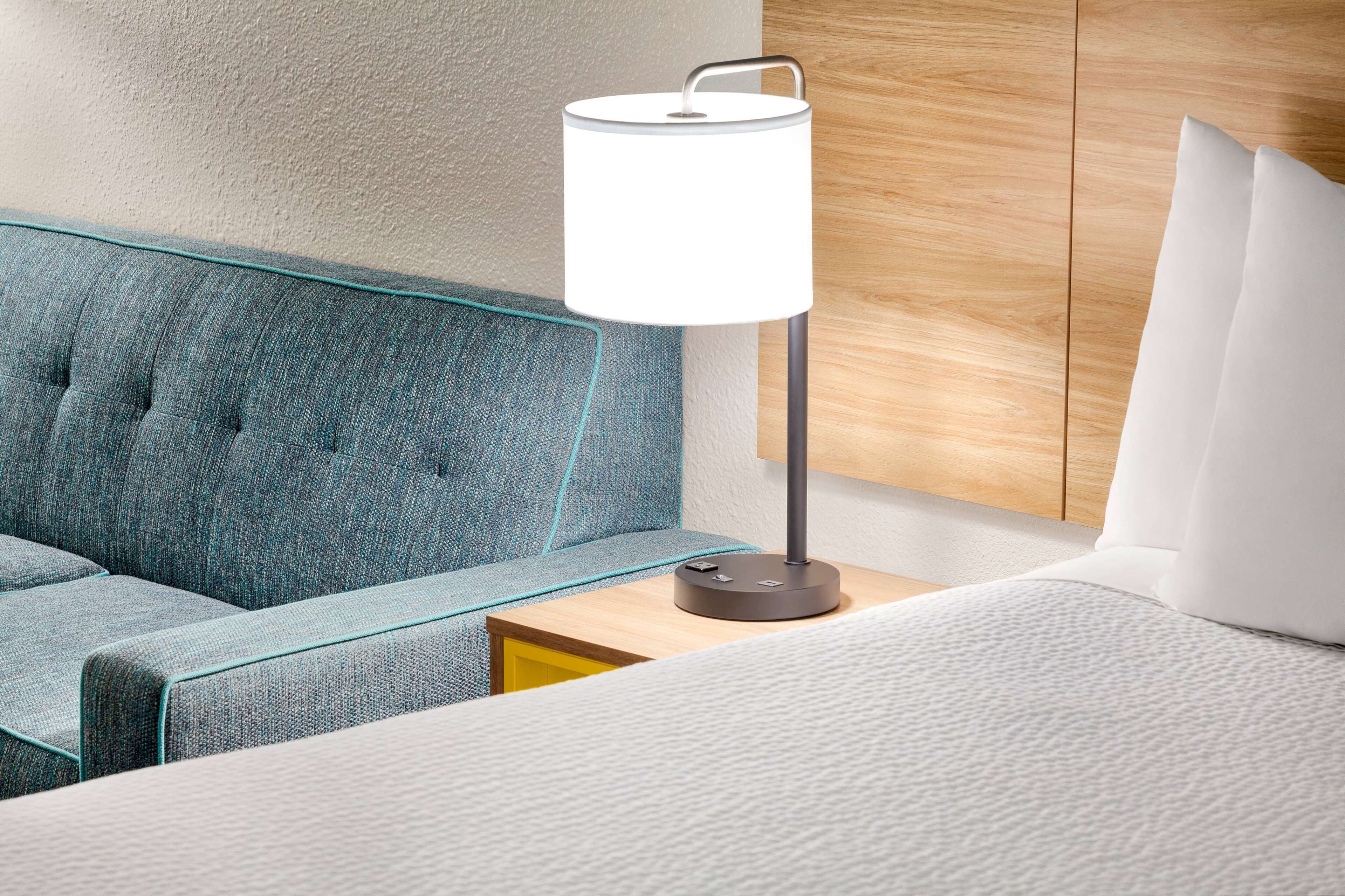 Ein modernes Hotelzimmer mit einem blau gepolsterten Sofa, einem Nachttisch mit Lampe und einem hölzernen Kopfteil.