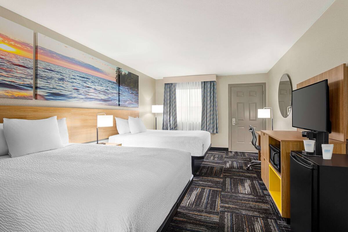 A hotel room with two beds, a TV, a desk with a coffee cup, and a painting of a sunset over the ocean.