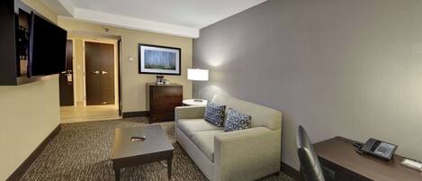 A hotel room with a sofa, a coffee table, a desk with a phone, a TV mounted on the wall, and a lamp.