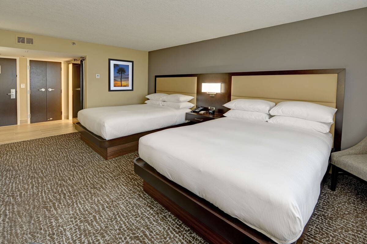 A hotel room with two beds, a nightstand, a lamp, and a door leading to another room.
