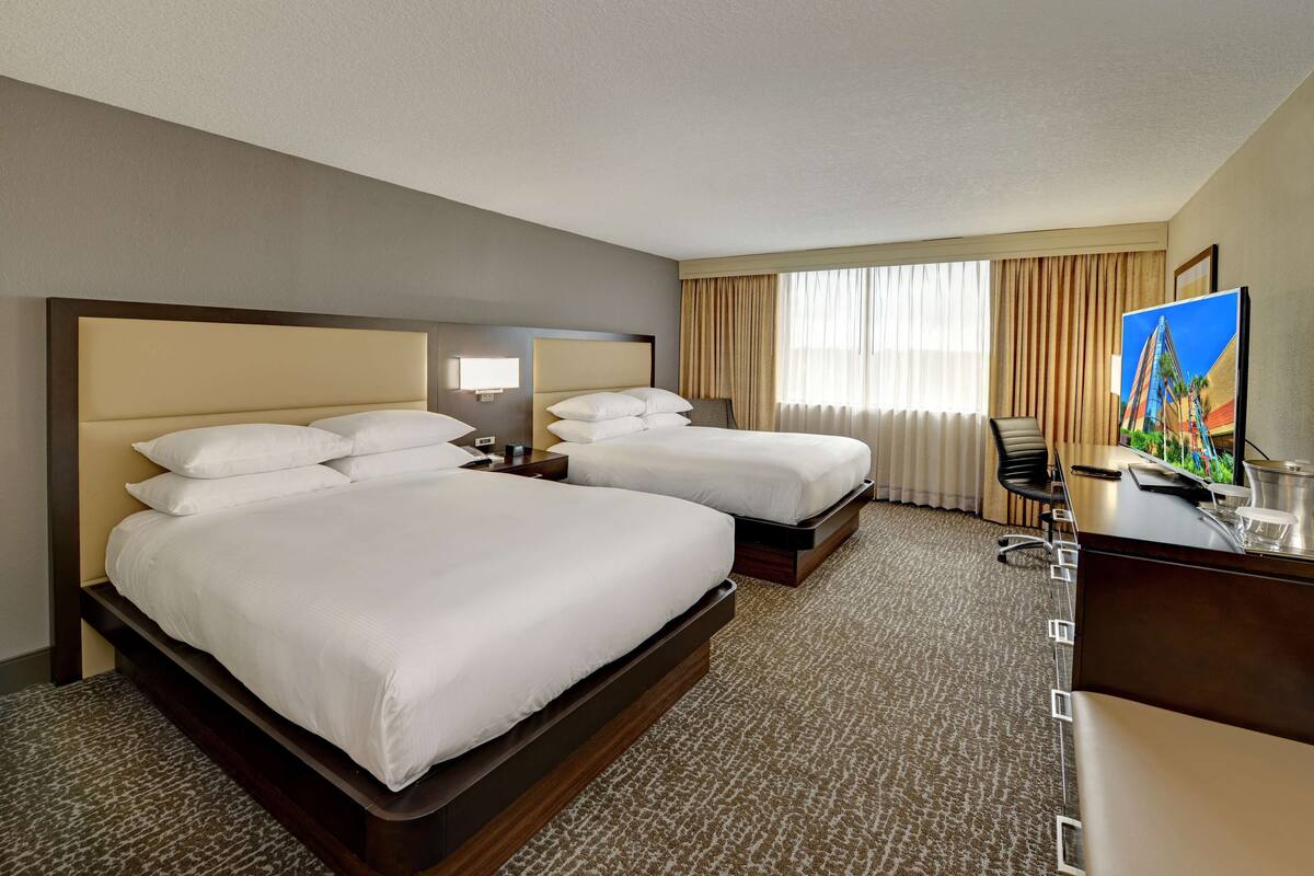A hotel room with two beds, a desk, a chair, a TV, and a large window with curtains.