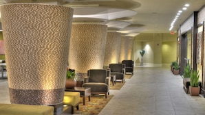 A hotel lobby with a long corridor, large textured pillars, seating areas, and potted plants.
