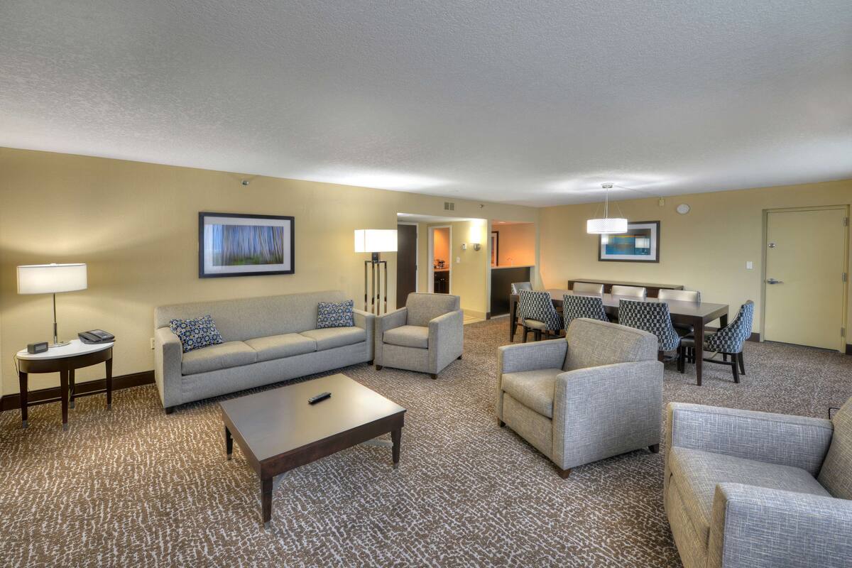 A hotel room with a living area featuring a sofa, armchairs, a coffee table, and a dining table with chairs.