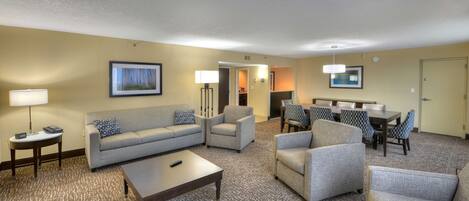 Habitación de hotel con sala de estar que incluye un sofá, sillones, una mesita de centro y una mesa de comedor con sillas.