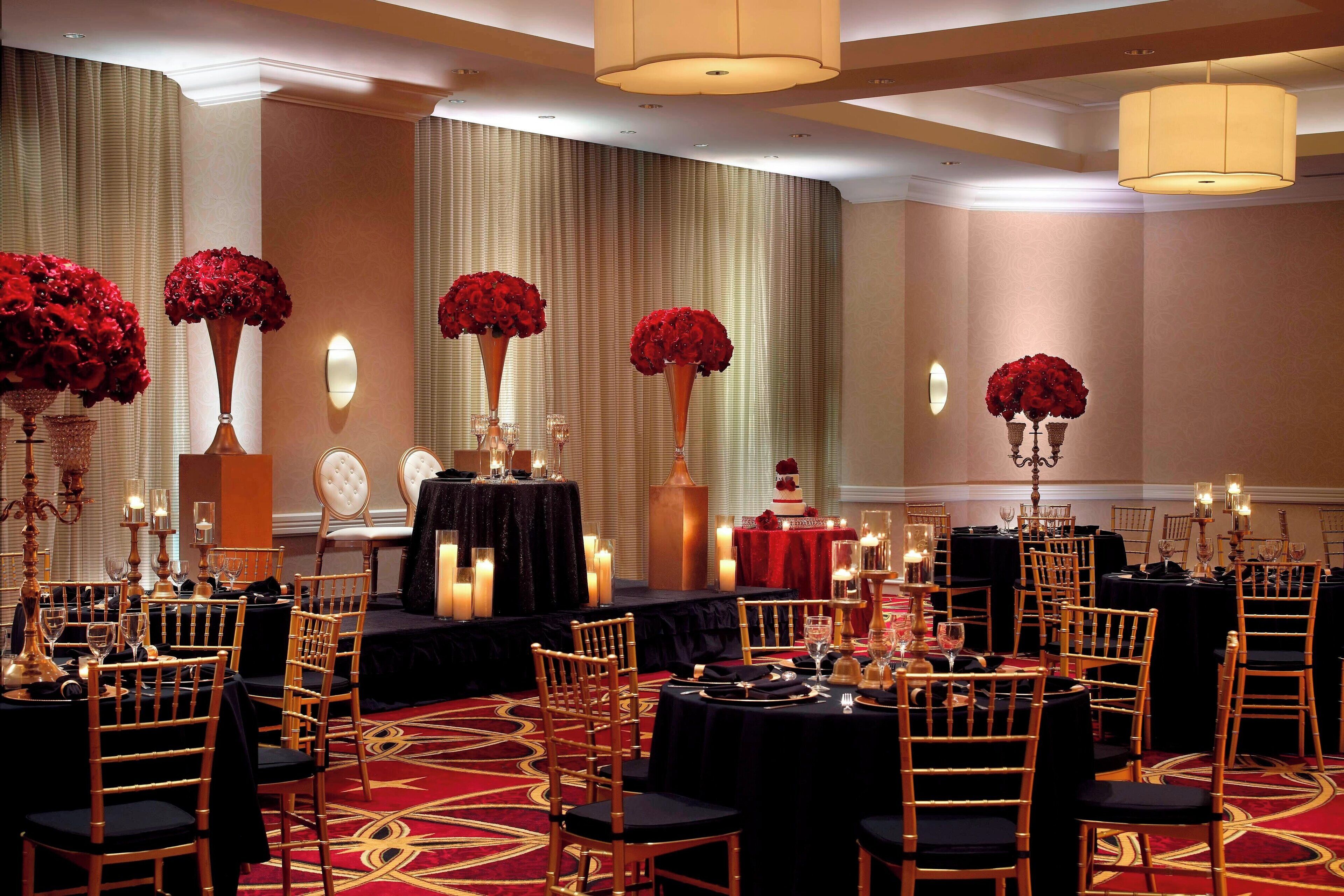 Un salón de banquetes con mesas redondas, manteles negros y centros de mesa florales rojos.