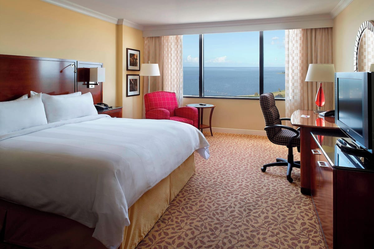 Habitación de hotel con una cama grande, un escritorio con televisor, una silla roja y vista al océano por la ventana.