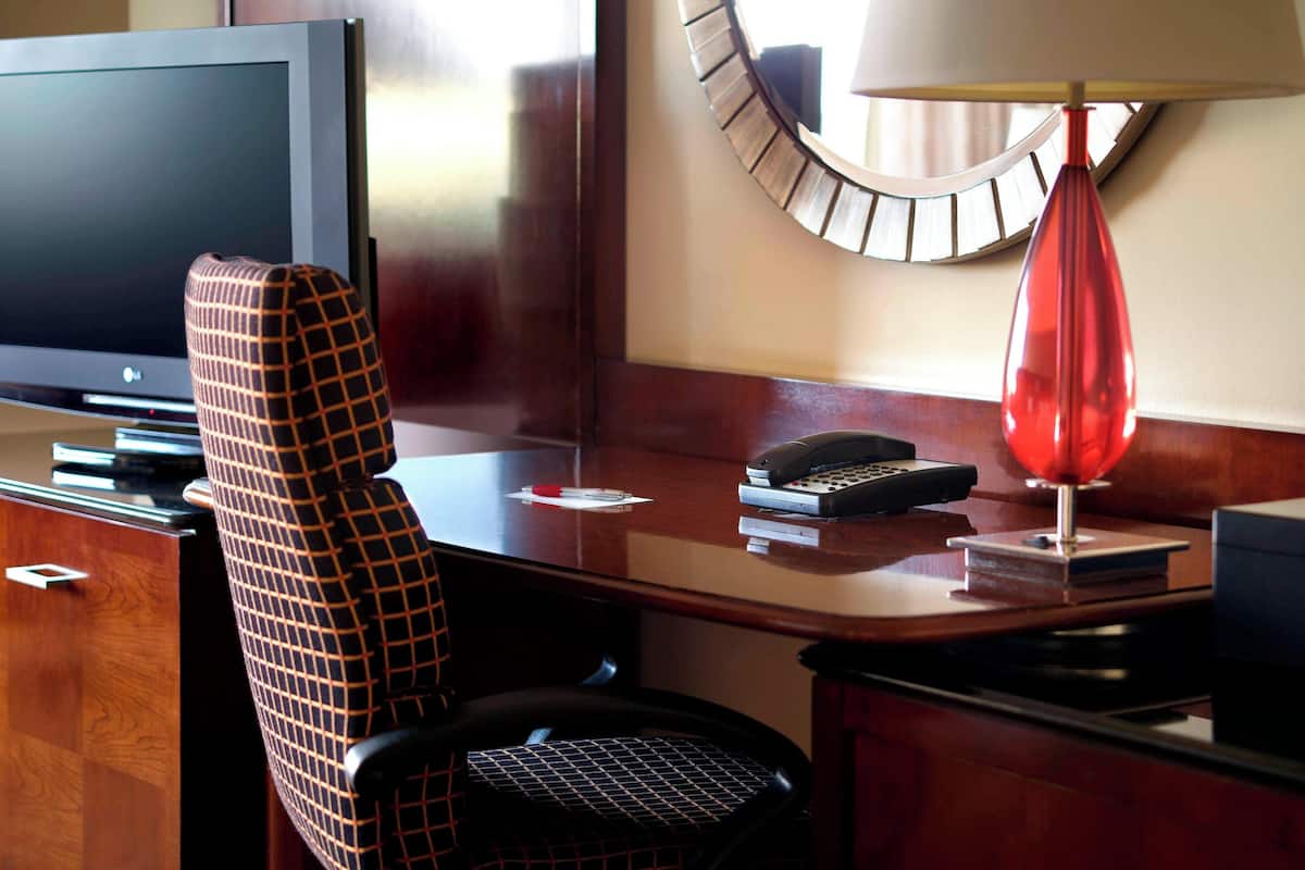 Habitación de hotel con televisión de pantalla plana, un sillón a cuadros, un escritorio con lámpara, teléfono y un reloj en la pared.