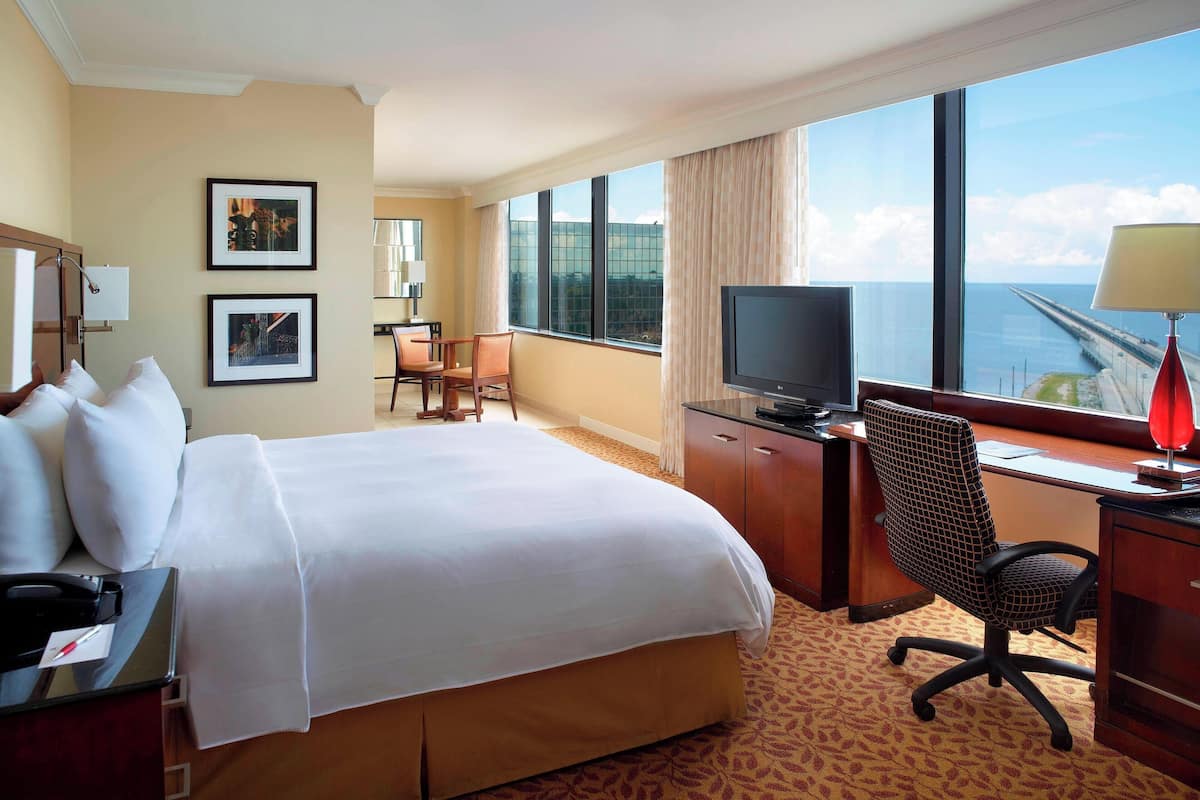 Habitación de hotel con una cama grande, un escritorio con computadora y televisor, una silla y vista al océano.