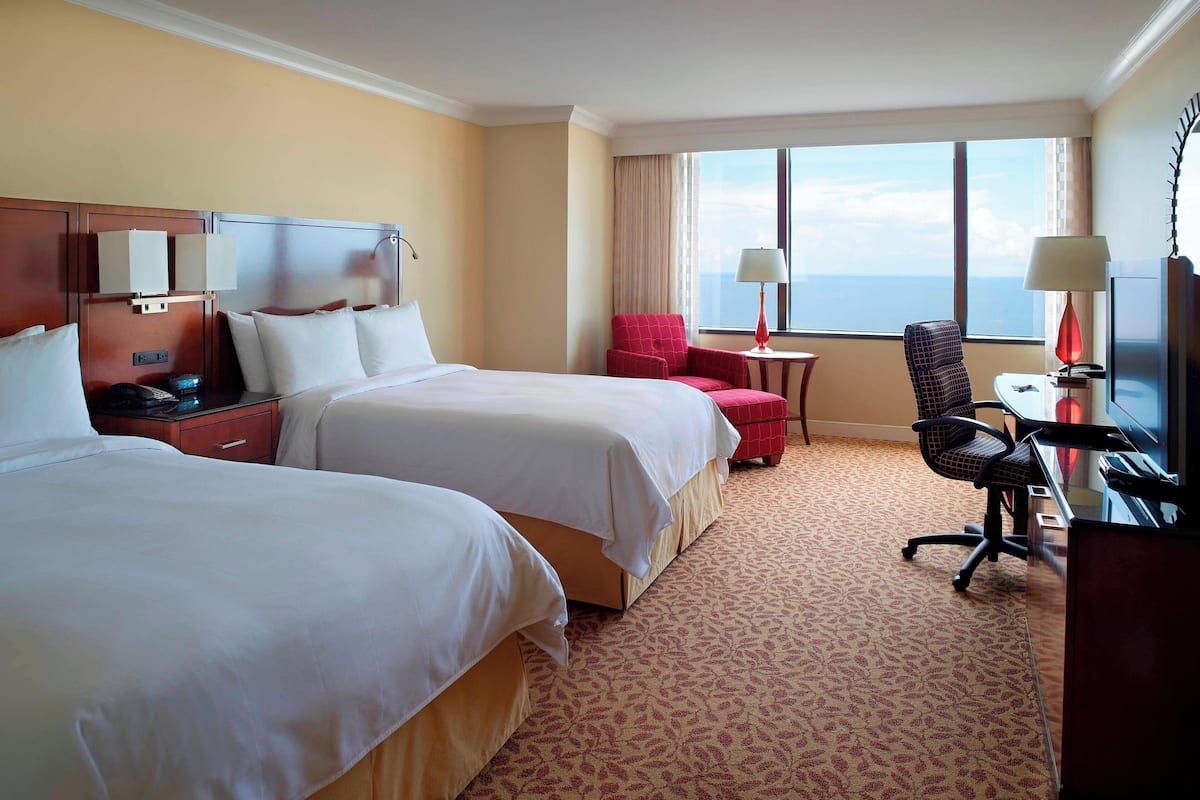Habitación de hotel con dos camas, un escritorio con silla, una mesita con lámpara y un ventanal con vistas.