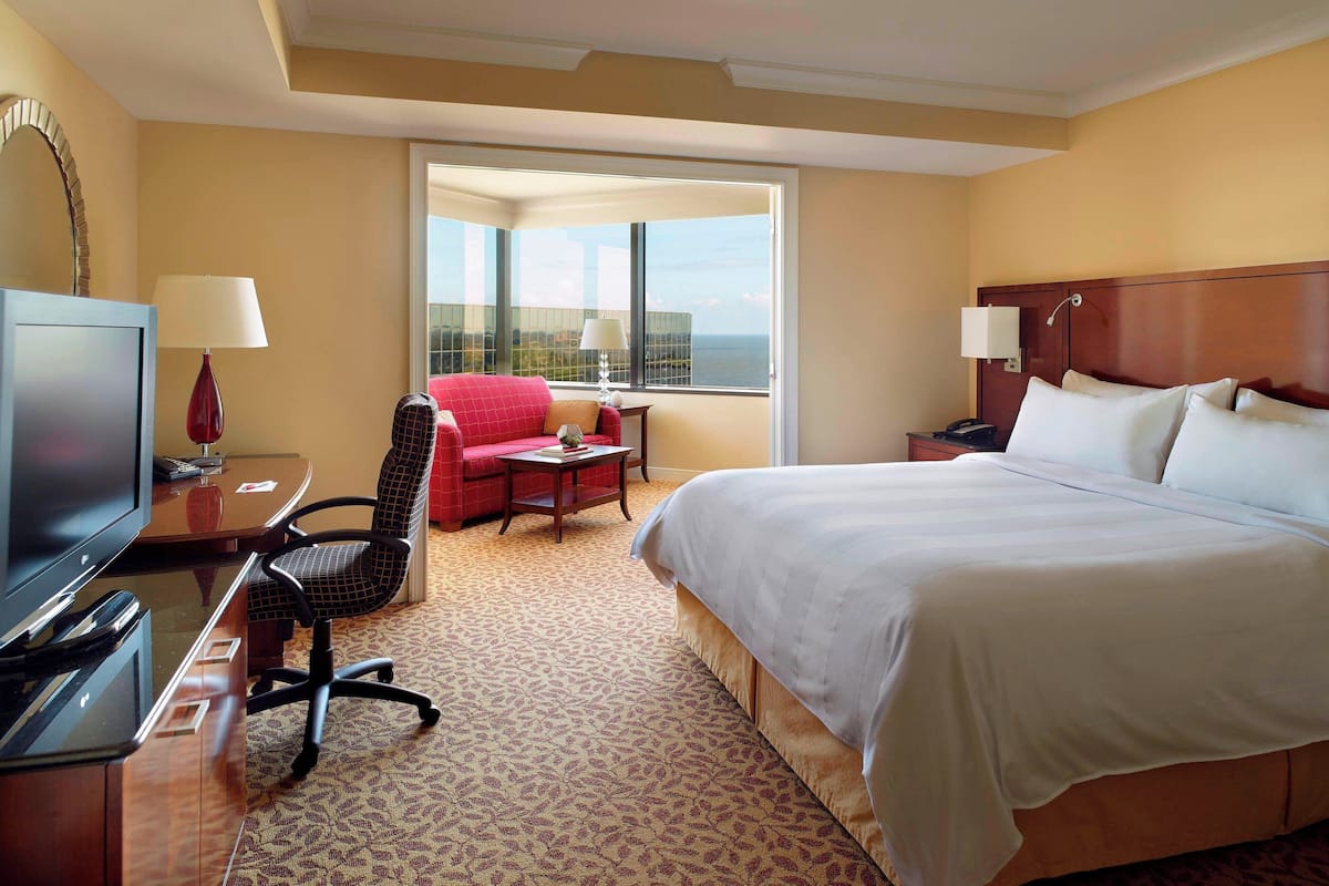 Habitación de hotel con una cama grande, un escritorio con televisor, un sillón rojo y vistas al océano a través de un ventanal amplio.