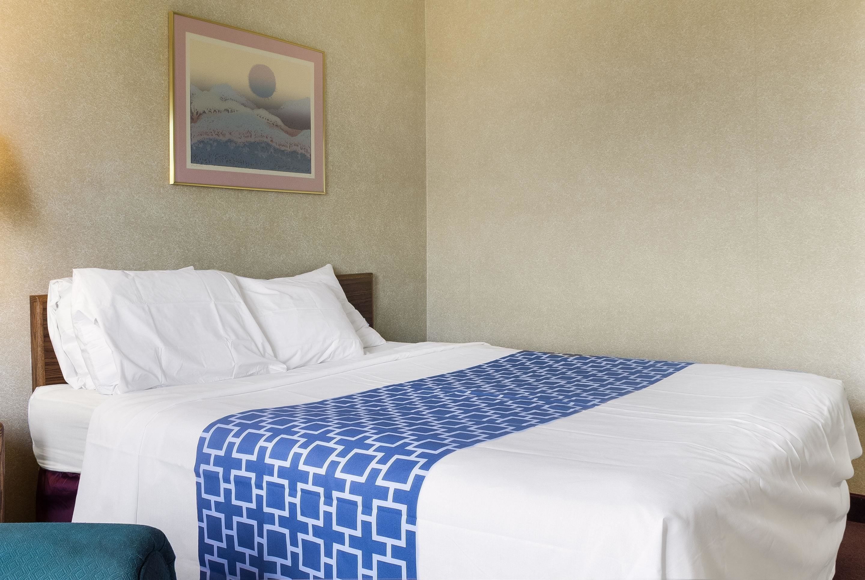 Een netjes opgemaakt bed met witte lakens en een blauw bedsprei, een ingelijst beeld aan de muur, en een teal-kleurige stoel op de voorgrond.