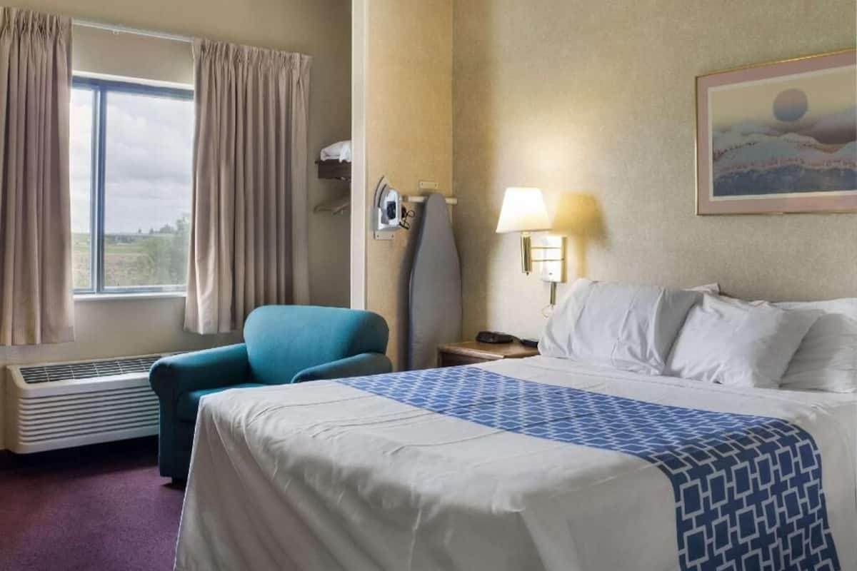 A hotel room with a bed, a chair, a window with curtains, a heater, a lamp, and a painting on the wall.