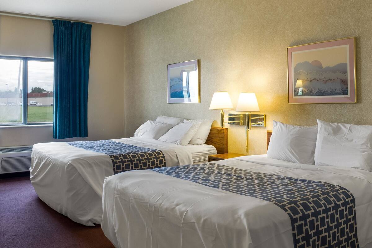 A hotel room with two beds, a large window with curtains, a wall-mounted lamp, and a framed picture.