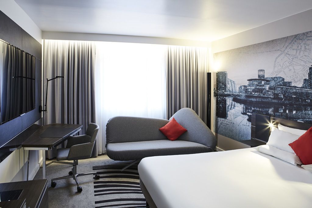 A hotel room with a bed, a desk, a chair, a TV, and a cityscape mural on the wall.
