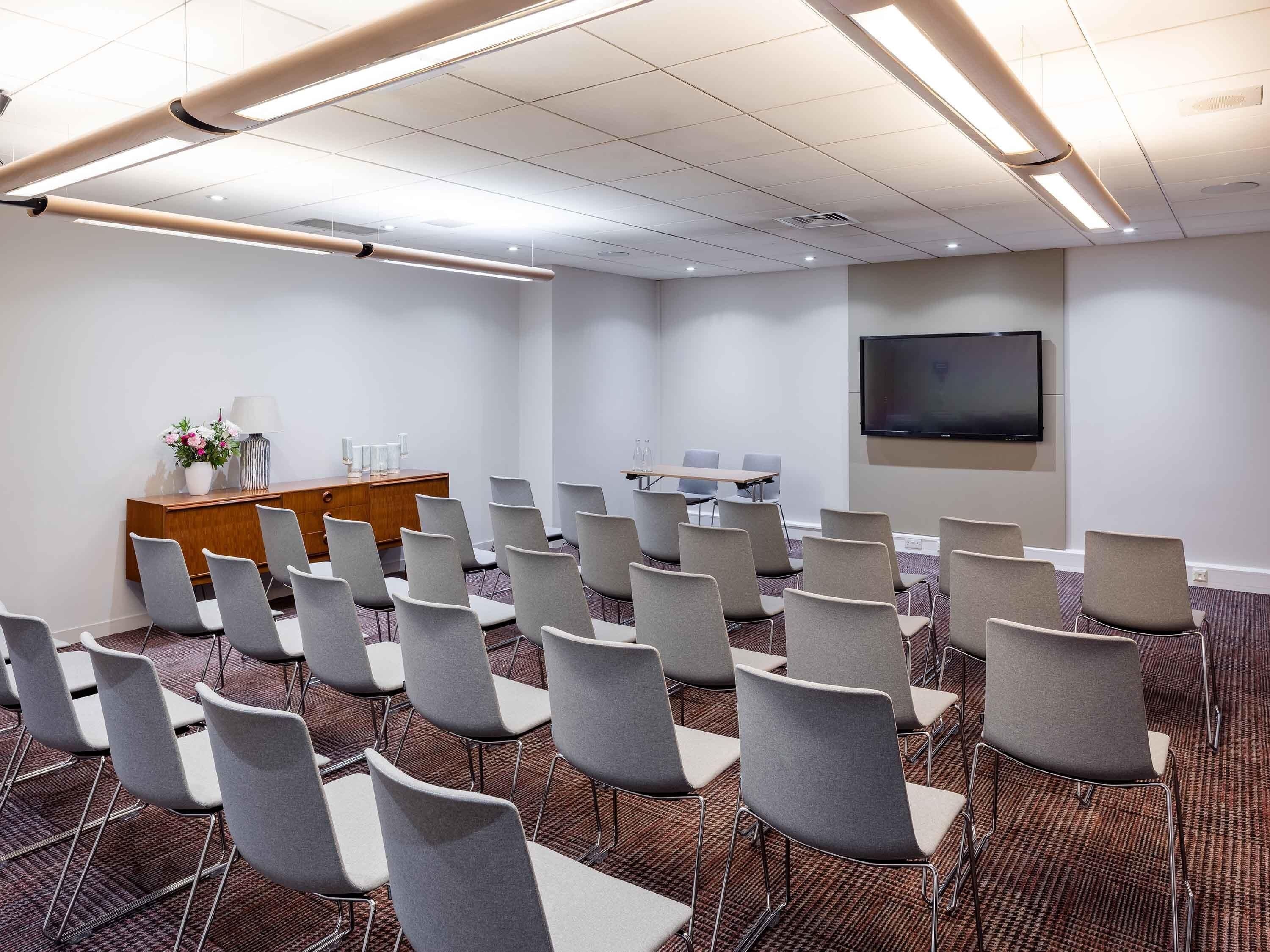 A conference room with rows of chairs, a wooden desk, a flat-screen TV, and a carpeted floor.
