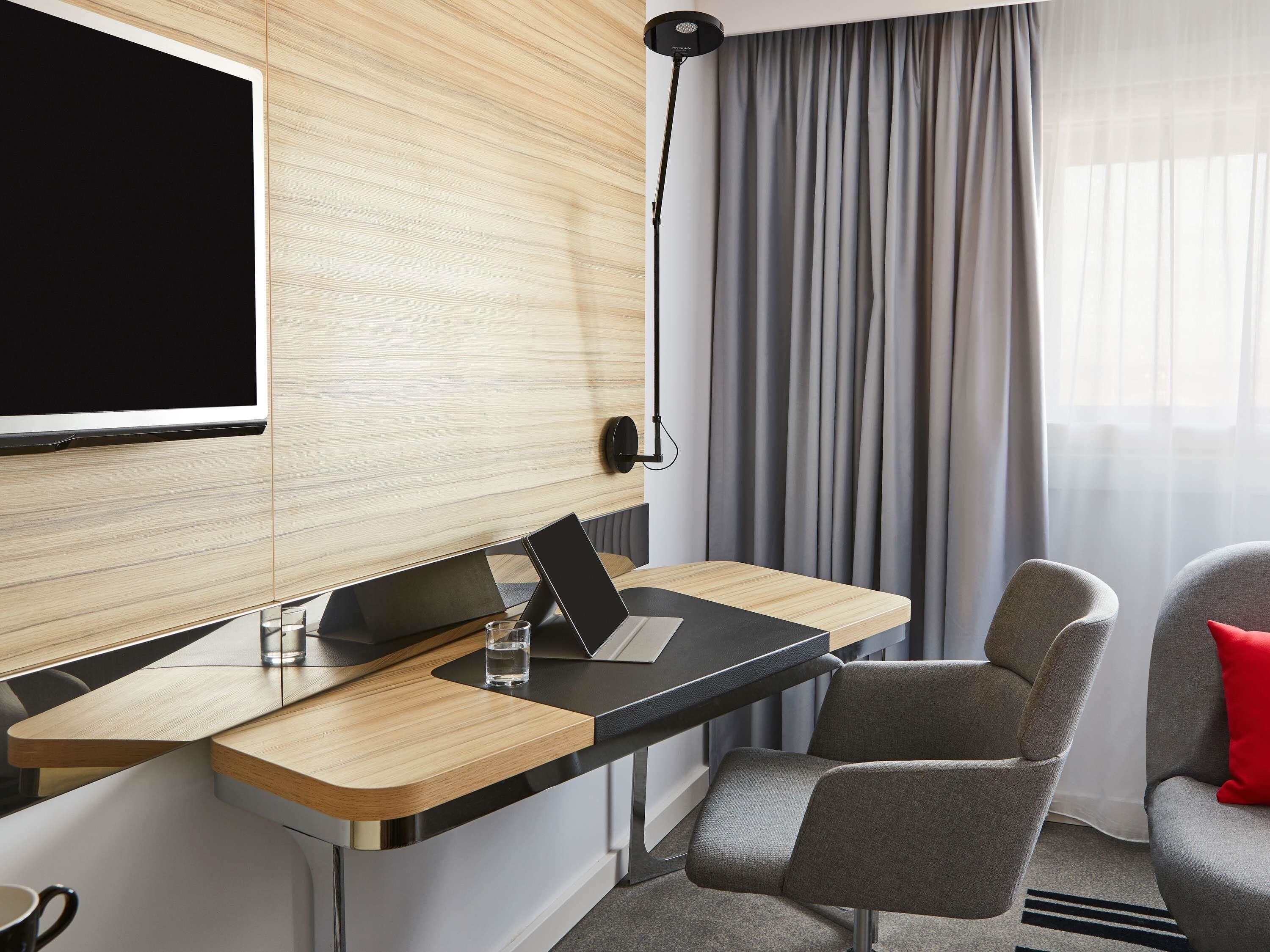 Een moderne hotelkamer met een houten accentmuur, een flatscreen televisie, een bureau met een laptop, een stoel en een rode kussen.