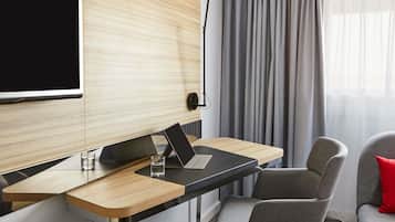 A modern hotel room with a wooden accent wall, a flat-screen TV, a desk with a laptop, a chair, and a red pillow.