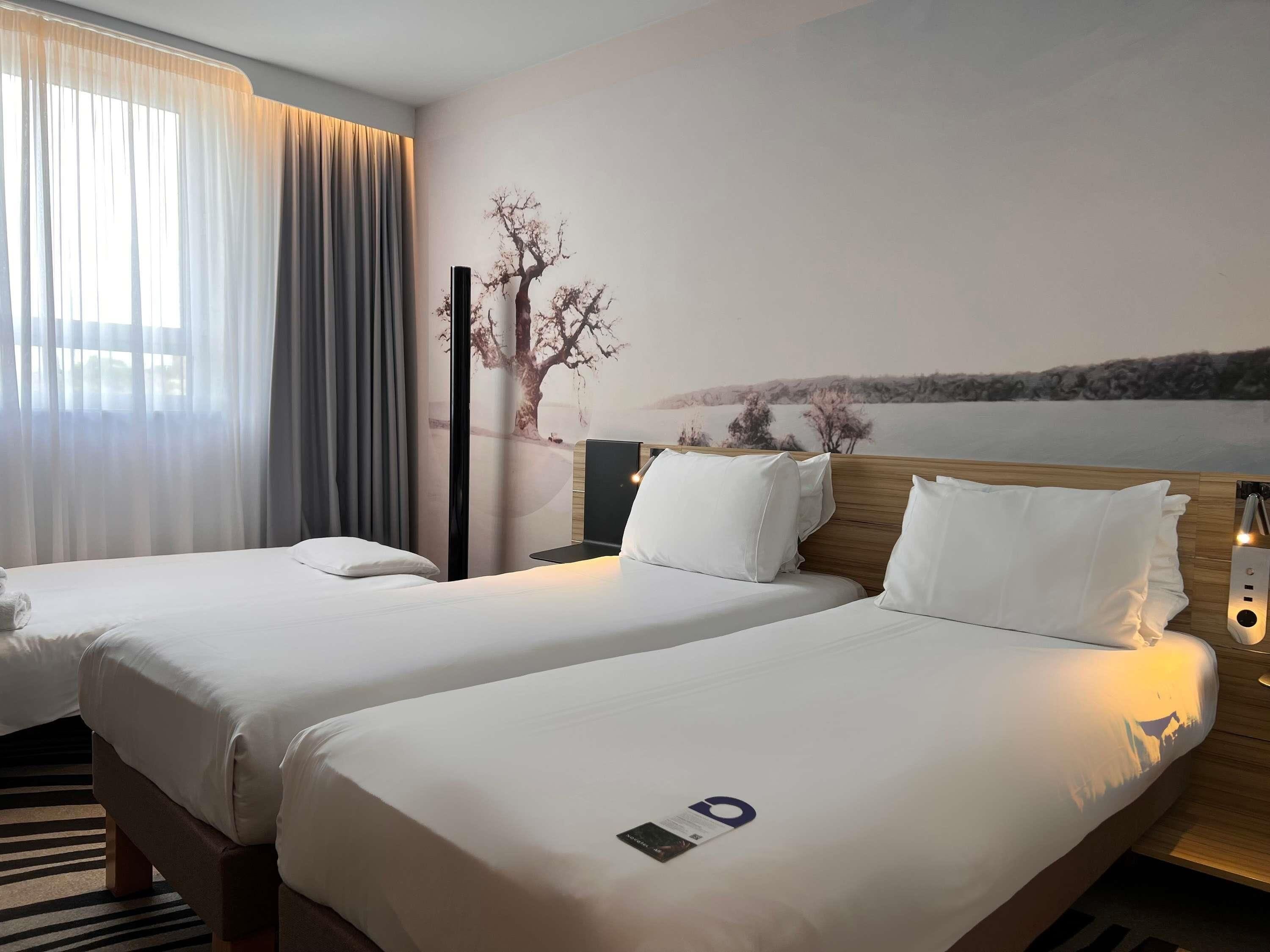 A hotel room with two beds, a nightstand with a lamp, a wall-mounted TV, and a scenic mural.