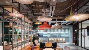 Ein modernes Interieur mit einer zentralen Sitzecke, Regalen mit Gegenständen und hängenden Pendelleuchten.