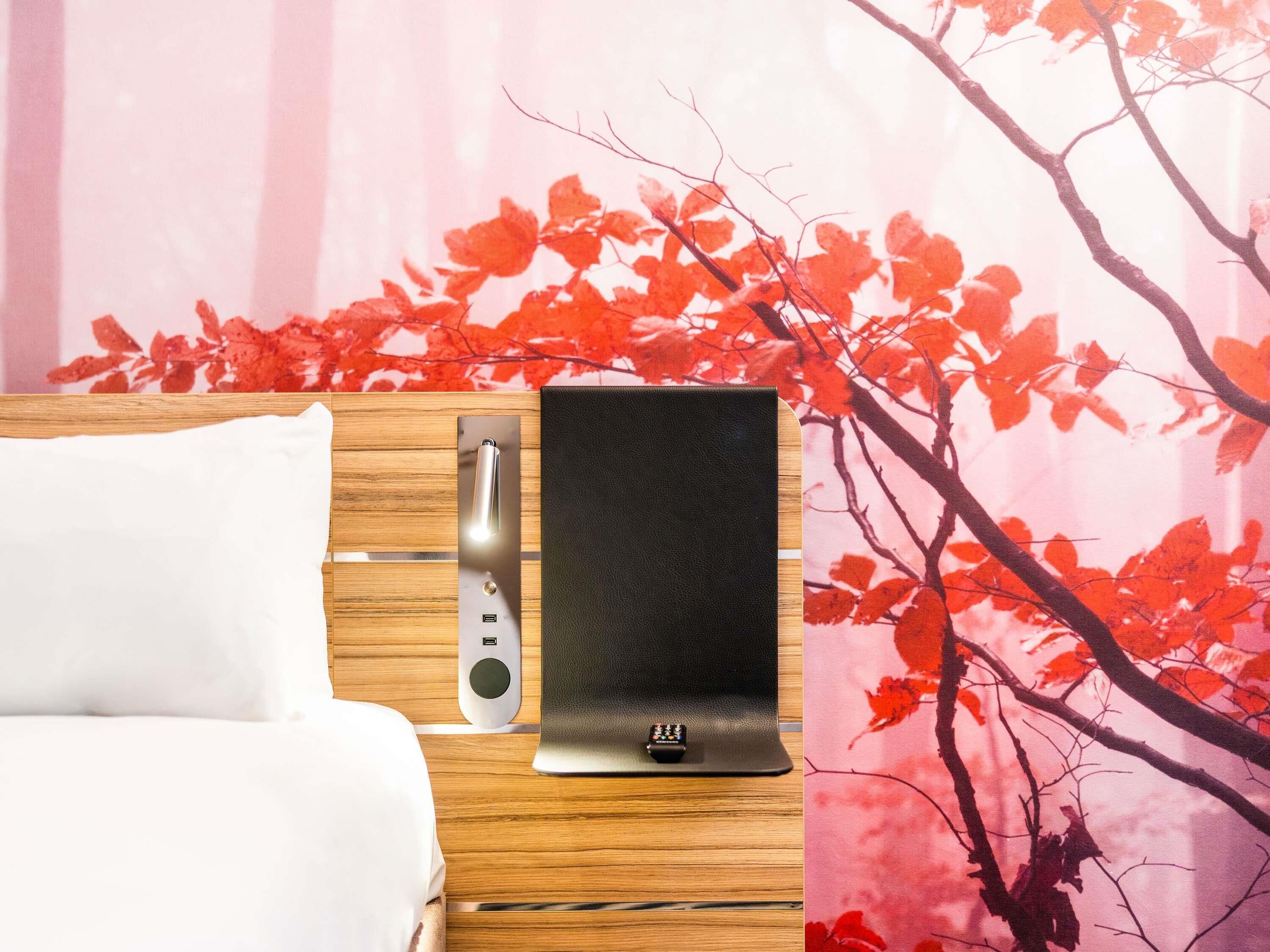 Ein Bett mit weißer Bettwäsche, einem Holz-Kopfteil, einem schwarzen elektronischen Gerät und einer Wand mit einem roten Blattmuster.