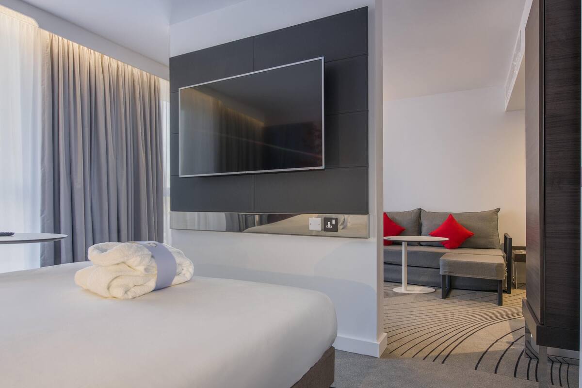 A modern hotel room with a large bed, a flat-screen TV mounted on the wall, a small table, and a sofa with red cushions.