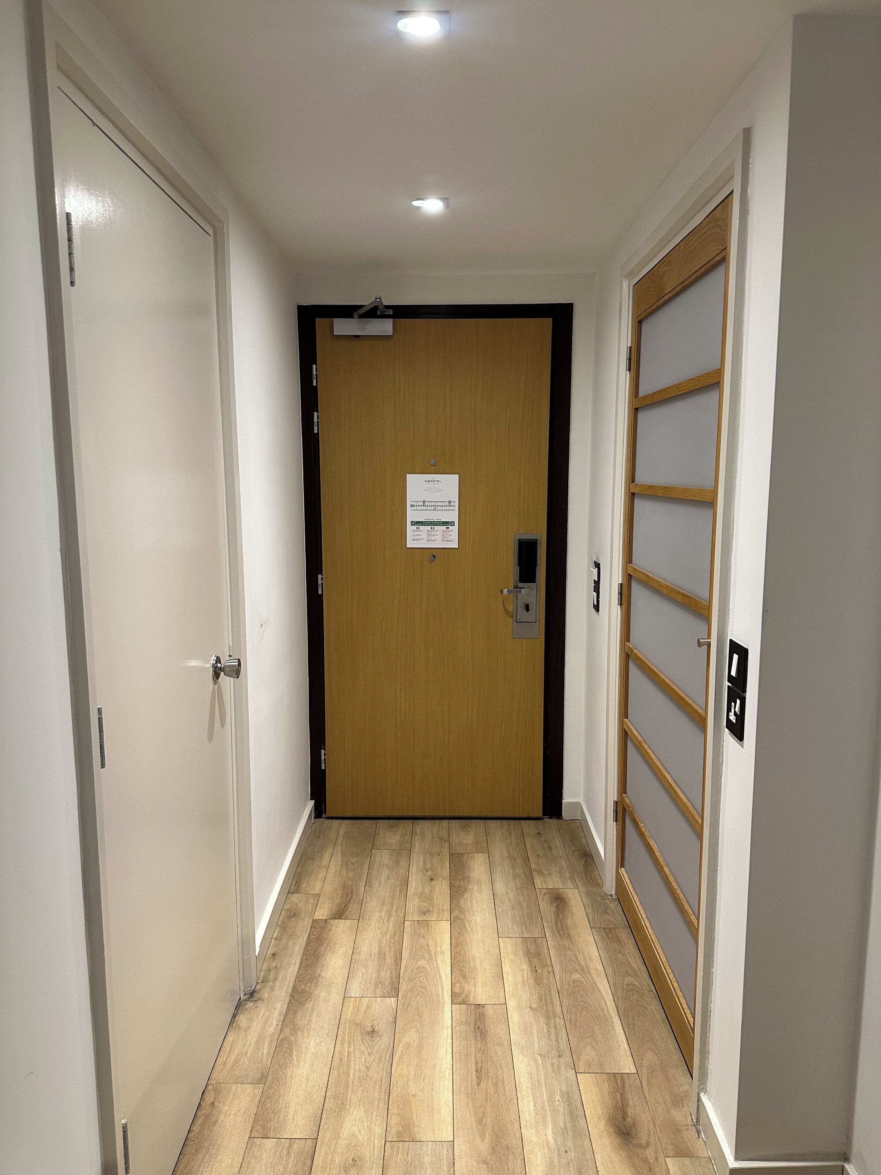 A hallway with a wooden door at the end, a light switch, and a fire alarm panel.