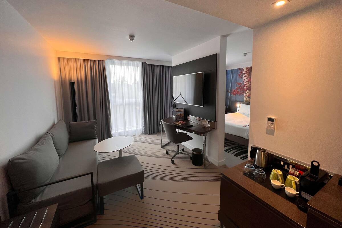 Une chambre d’hôtel équipée d’un canapé, d’une petite table, d’un bureau, d’un lit, d’une télévision, d’une machine à café et d’une fenêtre avec des rideaux.