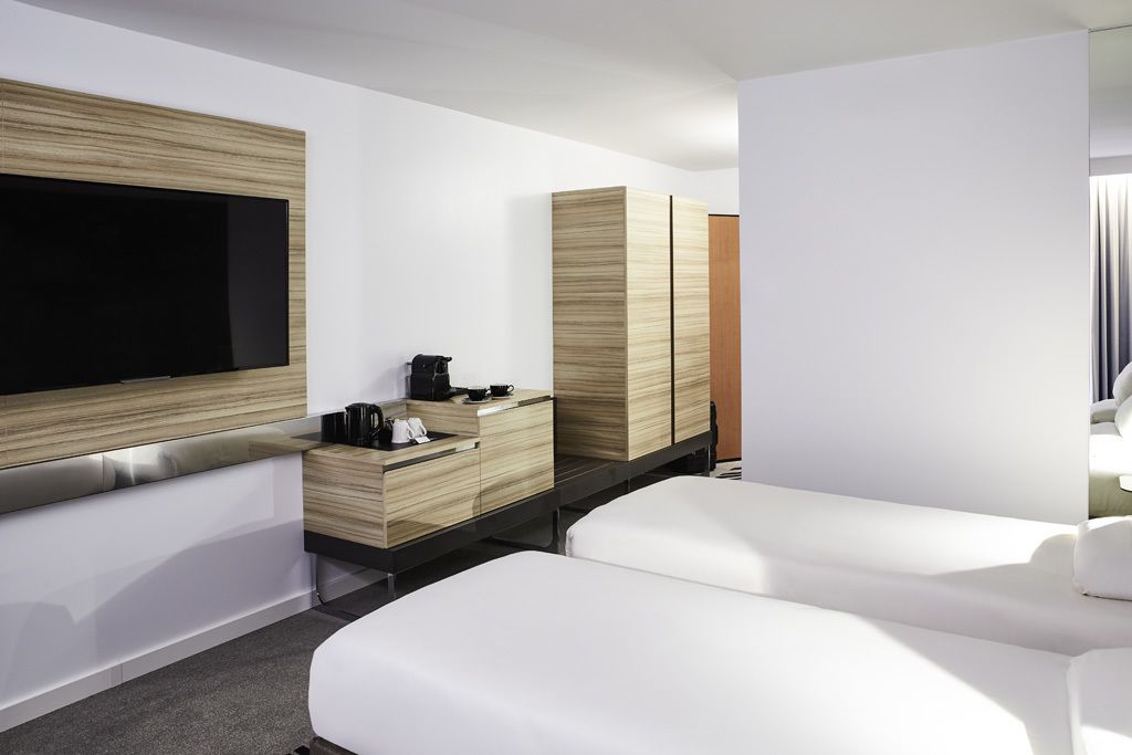 Quarto de hotel moderno com uma televisão de tela plana grande, um armário de madeira com cafeteira e duas camas brancas.