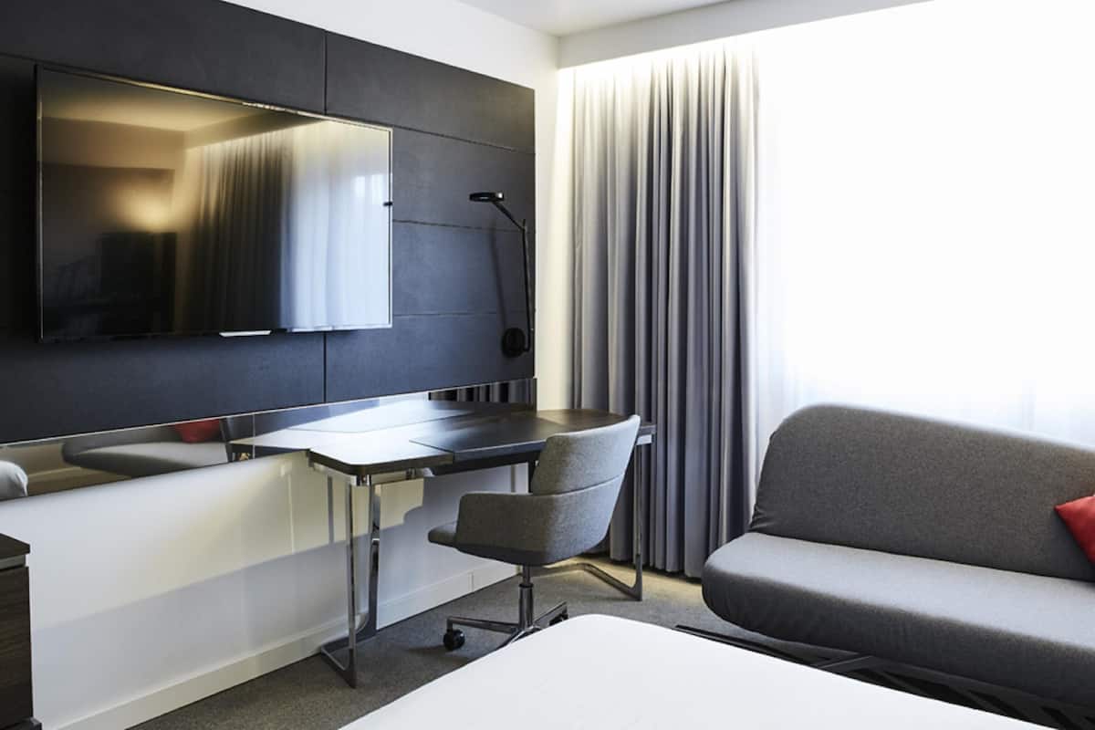 Una habitación de hotel moderna con televisión de pantalla plana, escritorio, silla, sofá y una ventana grande con cortinas.